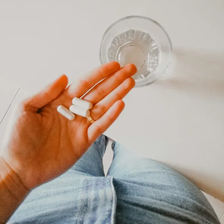 A hand holding three white capsules next to a glass of water