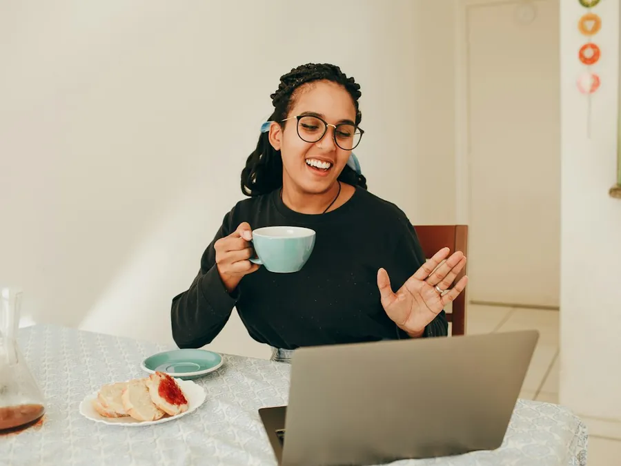 A woman on a video call at her laptop, holding a mug and smiling