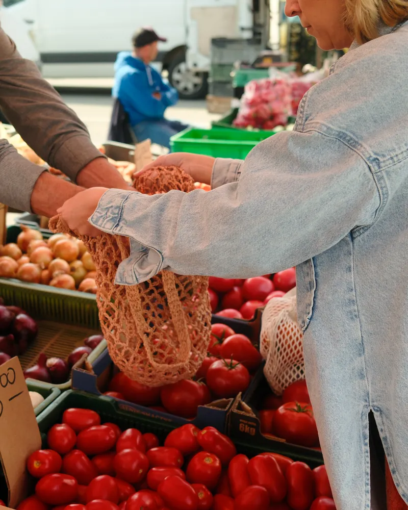 A shopper filling a mesh produce bag with tomatoes at a farmers market